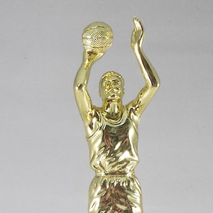 May include: A gold-colored basketball trophy featuring a player in a shooting pose, holding a basketball above their head. The trophy is mounted on a white marble base, showcasing a classic sports award design.