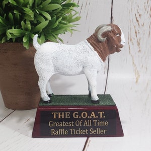 May include: A white and brown goat figurine with curved horns, standing on a green base. The base is mounted on a maroon plaque with gold lettering that reads "THE G.O.A.T. Greatest Of All Time Raffle Ticket Seller."