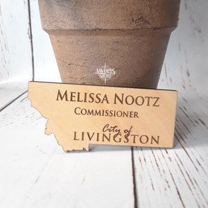 May include: A wooden name tag with the name "MELISSA NOOTZ" and the title "COMMISSIONER" engraved on it. The name tag is shaped like the state of Montana and includes the text "City of LIVINGSTON".