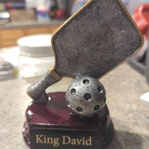 以下が含まれることがあります： 「King David」の文字が刻まれた台座に、パドルとボールが配置されたピクルボールのトロフィー。 シルバーとゴールドの色使いで、台座はマルーン色です。