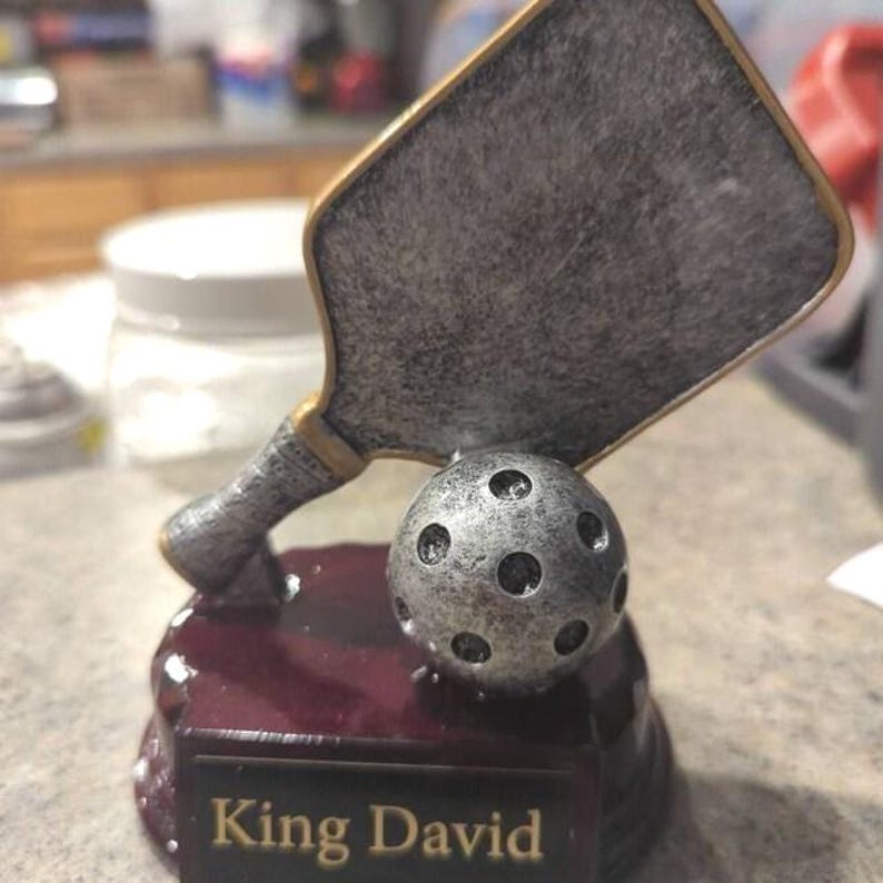 以下が含まれることがあります： 「King David」の文字が刻まれた台座に、パドルとボールが配置されたピクルボールのトロフィー。 シルバーとゴールドの色使いで、台座はマルーン色です。