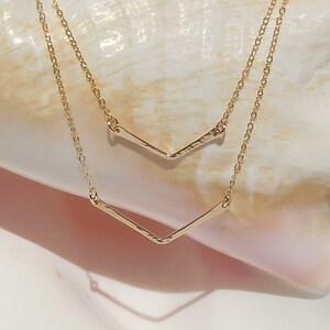 Puede incluir: Un collar de oro con dos cadenas delicadas y un colgante de chevron. El collar se muestra sobre una concha.