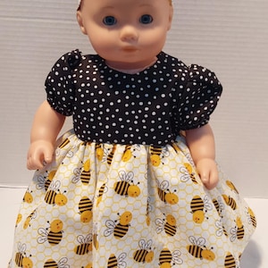 May include: A doll wearing a black and white polka dot top with a yellow and white bee-themed skirt. The doll also has a matching headband. The skirt features a honeycomb pattern with yellow and black bees.