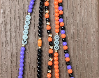 Puede incluir: Un llavero con temática de Halloween con múltiples hebras de cuentas en morado, negro y naranja. Las hebras presentan las palabras "WITCHY" y "BOO" deletreadas en cuentas, junto con amuletos de una bruja en una escoba, un murciélago, un gato y una calabaza.
