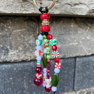 May include: A festive keychain with a gold ring and a beaded tassel. The tassel features red, green, white, and clear beads, including candy cane and Santa Claus designs. The keychain is a holiday-themed accessory.