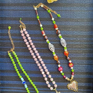 May include: Four beaded necklaces with gold-tone accents and pendants. One necklace is green with a gold charm, another is pink with a flower pendant, and the third is multi-colored with a heart pendant. The necklaces are on a blue striped background.