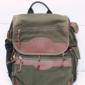 May include: A green canvas backpack with brown leather accents. The backpack features multiple zippered compartments, leather zipper pulls, and the "ORVIS" logo. A mesh pocket is visible on the side.