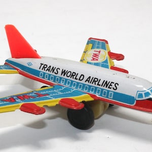 May include: A vintage toy airplane with the text "TRANS WORLD AIRLINES" printed on the white fuselage. The wings are blue and yellow with red accents. The tail fin is red. The plane has a black nose and a single black wheel.