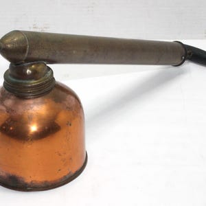 May include: An antique copper and brass oil can with a long spout and a black handle. The oil can has a rounded base and a cylindrical spout, showing signs of age and use. The metal has a warm, aged patina.