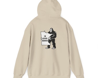 May include: A beige hoodie with a hood and long sleeves. The back features a black and white graphic of a Bigfoot holding a sign that reads "IN GOOD HANDS". The hoodie is made of a soft, comfortable material.