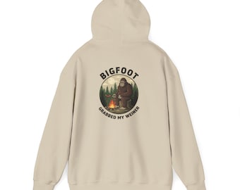 May include: Beige hoodie with a Bigfoot graphic on the back. The graphic features Bigfoot sitting by a campfire with the text "BIGFOOT GRABBED MY WEINER" in a circular design. The hoodie has a hood and a kangaroo pocket.