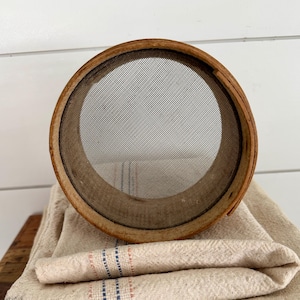May include: A wooden sieve with a fine mesh screen rests on a stack of folded, cream-coloured linen towels. The towels feature subtle stripes in blue, red, and grey. The sieve's wooden frame shows signs of age, adding to its rustic charm.