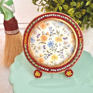 May include: A circular picture frame featuring a red mosaic border with small green and yellow floral accents. The frame stands on two small, matching mosaic feet and contains a paper insert decorated with a pattern of yellow and blue flowers.