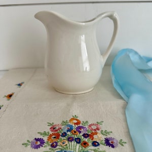 May include: A white ceramic creamer with a floral embroidered linen napkin and a blue ribbon. The embroidery features a bouquet of colorful flowers with a blue ribbon.