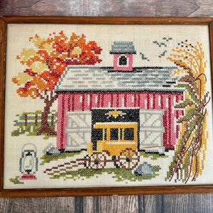 May include: Framed cross-stitch artwork depicting a red barn with a gray roof, a yellow wagon, and an autumn tree. The artwork is set against a beige background and framed in a wooden border. The scene includes a fence, a lantern, and a bundle of wheat.