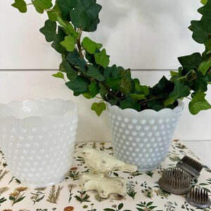 May include: Two white milk glass bud vases with a scalloped rim and a textured, dotted surface. The vases are filled with green ivy and are sitting on a floral patterned surface. There is a small white bird figurine on the surface.