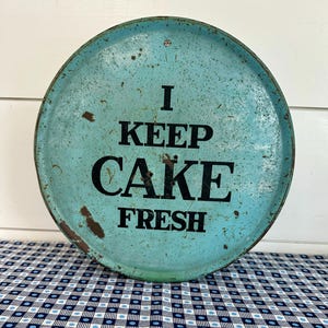 May include: A round, distressed, turquoise metal cake plate with the words "I KEEP CAKE FRESH" in black lettering. The plate has a rusty rim and is sitting on a blue and white checkered tablecloth.