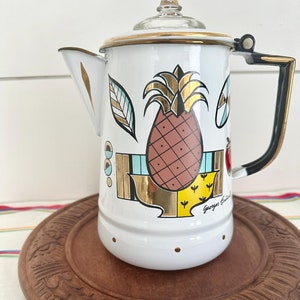 May include: White enamel coffee pot with a black handle and a pineapple design. The design features a pineapple with gold accents, a leaf, and geometric shapes. The pot has a gold rim and a glass lid. The text "George's Britain" is visible on the pot.