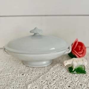 May include: A white porcelain serving dish with a lid and two handles. The dish is oval shaped and has a raised center. The dish is sitting on a white crocheted tablecloth.
