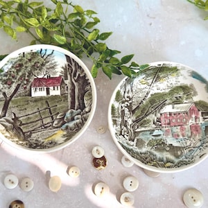 May include: Two round ceramic dishes featuring detailed landscape illustrations. The left dish depicts a white house with a red roof near a wooden fence and trees. The right dish shows a red building by a body of water with surrounding trees.