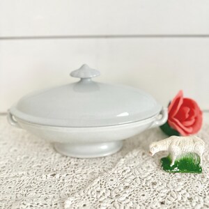 May include: A pale grey ceramic serving dish with a lid, handles, and a pedestal base. A red rose and a small white lamb figurine with green grass are next to the dish. The dish sits on a white lace tablecloth.
