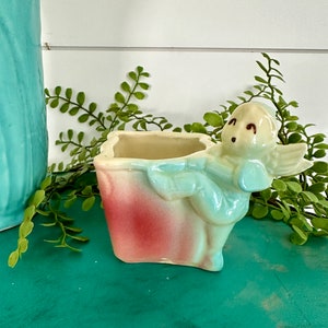 May include: A vintage ceramic planter shaped like a girl holding a pot. The planter is painted in shades of pink, blue, and white.