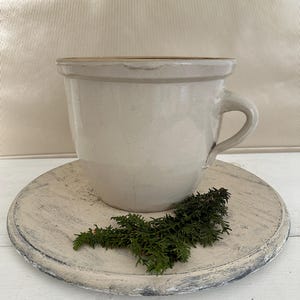 May include: A white ceramic crock with a handle, sitting on a white wooden round platter with green sprigs of greenery.