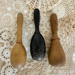 May include: Three antique wooden scoops with handles. One is painted black, and the other two are natural wood tones. The scoops are displayed on a cream-colored, open-weave textile.