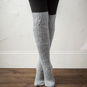 May include: A pair of gray knee-high socks with a cable knit pattern. The socks are worn over black leggings.