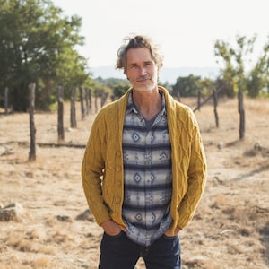 Puede incluir: Un hombre con un cárdigan amarillo sobre una camisa de estampado azul y blanco, de pie en un campo con hierba seca y postes de madera de una valla en el fondo.