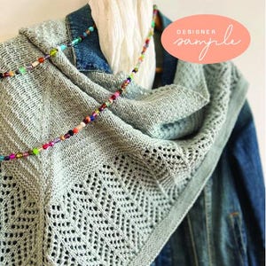 May include: A pale grey knitted shawl with a geometric lace design and a beaded necklace. The shawl is draped over a denim jacket. The text "DESIGNER sample" is in a peach oval.