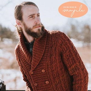 May include: A rust-colored, double-breasted cable-knit cardigan with large wooden buttons. The cardigan has a shawl collar and is labeled "DESIGNER sample" in an oval. The garment is worn over a plaid shirt.