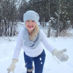 May include: A woman wearing a light blue knitted hat and scarf, white sweater, blue jeans, and brown boots is standing in the snow. She is holding a pair of white mittens in her hands.
