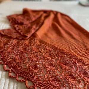 May include: A crocheted shawl in a burnt orange color with a lacy pattern. The shawl is draped over a white bedspread.