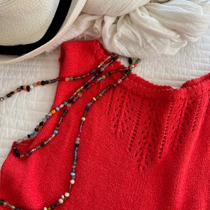 May include: A red sleeveless knitted top with a delicate openwork design around the neckline. A multi-colored beaded necklace is draped over the top.