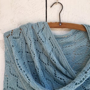 May include: A light blue crocheted shawl with a delicate openwork pattern hangs on a wooden hanger against a white wood background.