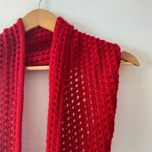 May include: A red crocheted scarf with a lacy pattern hangs on a wooden hanger.