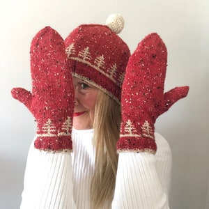 May include: A red knit hat with a white pom pom and a white knit pattern of trees. The hat is paired with matching red knit mittens with a white knit pattern of trees.