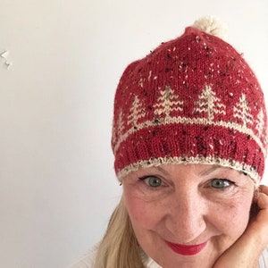 Puede incluir: Un gorro de punto rojo con un pompón blanco en la parte superior. El gorro tiene una banda de punto blanco con un patrón de pinos estilizados.