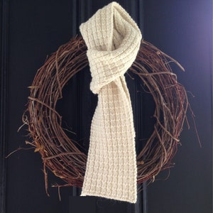 May include: A white knitted scarf is tied around a brown twig wreath. The wreath is hanging on a black door.