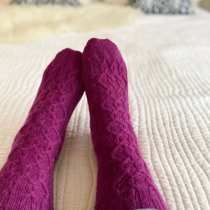 Peut inclure: Une paire de chaussettes en tricot violet avec un motif en câble. Les chaussettes sont portées par une personne assise sur un lit blanc.