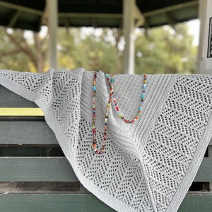 May include: A gray crocheted shawl with a delicate lace pattern draped over a wooden bench. A colorful beaded necklace is resting on top of the shawl.