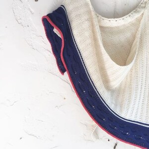 May include: Cream-colored knitted shawl with a navy blue border and a red trim. The shawl is hanging on a wooden hanger against a white textured wall. The knitted pattern is visible throughout the shawl.