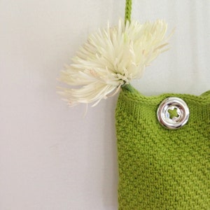 May include: A small green knitted bag with a silver button closure. A white flower is hanging from the strap of the bag.