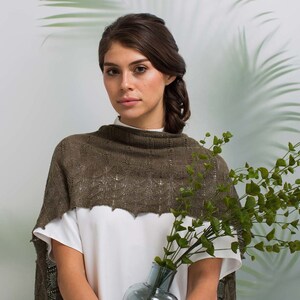 May include: A woman wearing a brown knitted shawl with a delicate lace pattern. She is standing in front of a white wall with green plants in the background.
