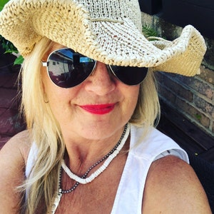 May include: A woman wearing a straw cowboy hat with a wide brim, sunglasses, and a white tank top with a layered necklace.