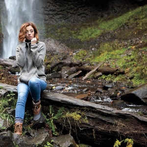 Puede incluir: Una mujer con un suéter gris y jeans azules se sienta en un tronco cerca de una cascada. Lleva botas marrones y tiene las manos en su suéter.