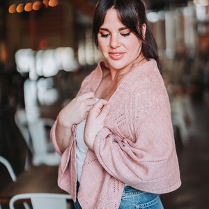 Può includere: Una donna indossa un cardigan di maglia rosa con un motivo delicato, una canottiera bianca e jeans blu. Il cardigan ha una vestibilità rilassata e maniche lunghe.