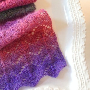 May include: A purple, pink, and red ombre knitted scarf with a lacy pattern. The scarf is folded and displayed on a white plate with a decorative edge.