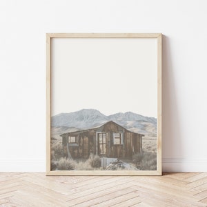 May include: A framed photograph of a rustic wooden cabin in a mountainous landscape. The cabin is weathered and has a single window. The mountains in the background are covered in snow.